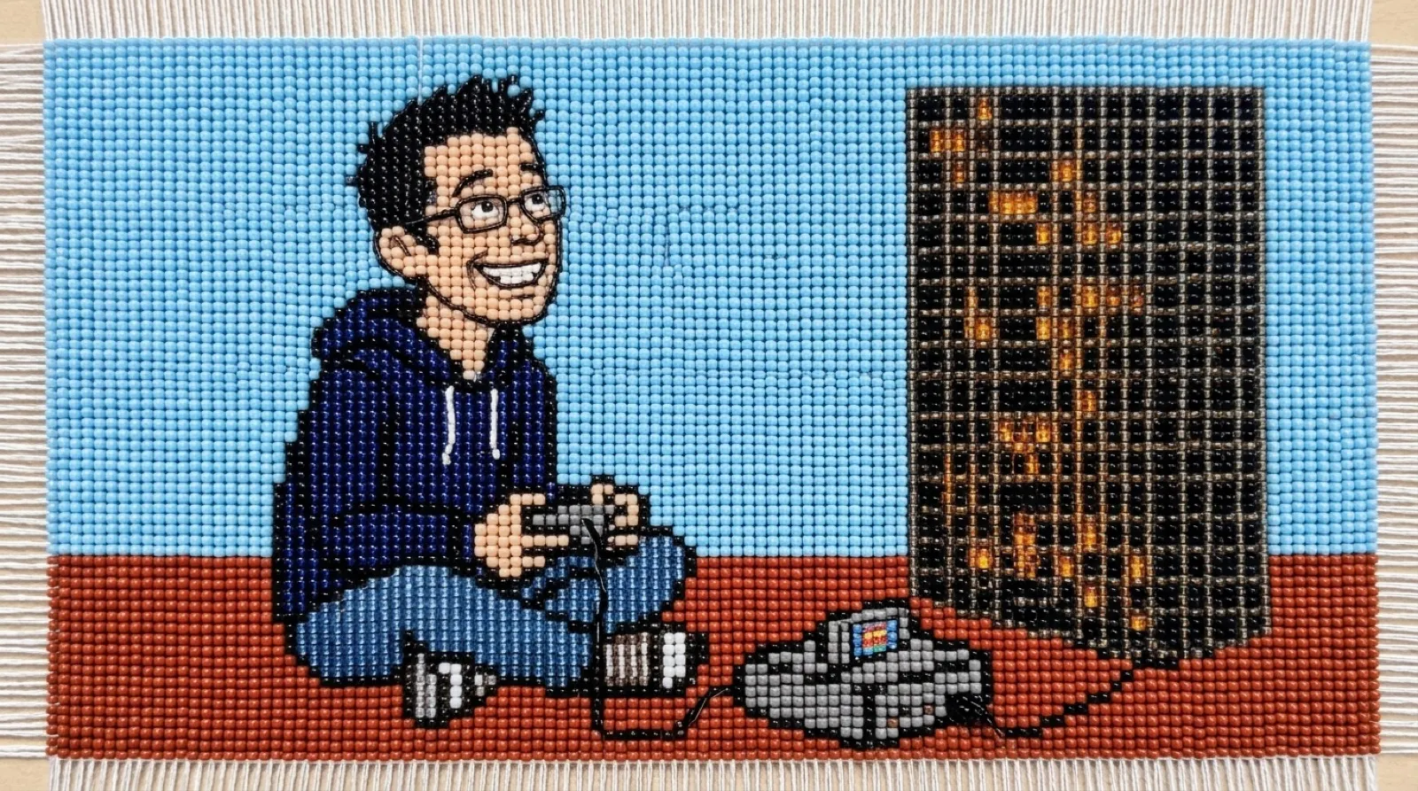 Chris Pirillo in glass-bead loom work, holding an old game controller beside a tall grid of tiny glowing cells and a boxy gray game console