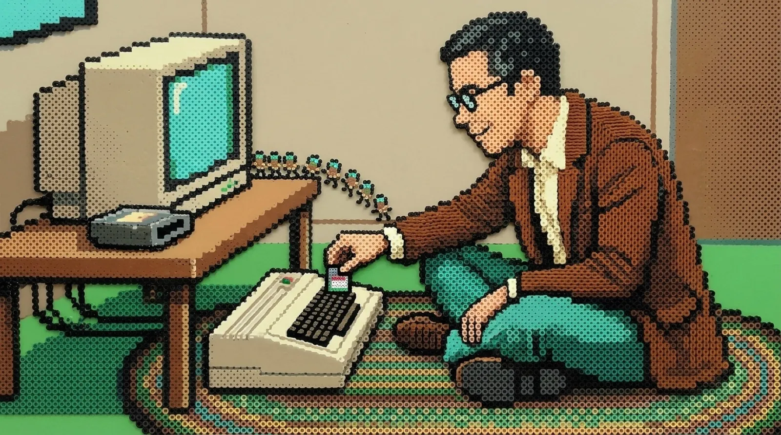 Chris, sitting cross-legged on the floor, pushes a tiny memory card into a cartridge plugged into an old beige home computer while a line of small marching creatures walks off its screen, all made of ironed plastic fuse beads.